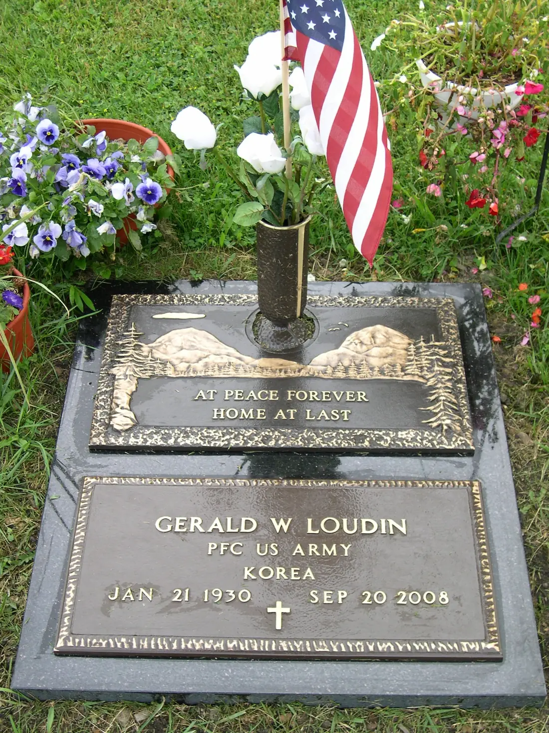 A bronze marker on a granite base giving a private first class's name, branch and war, with a cross emblem, a flower vase, planted flowers and a small American flag.